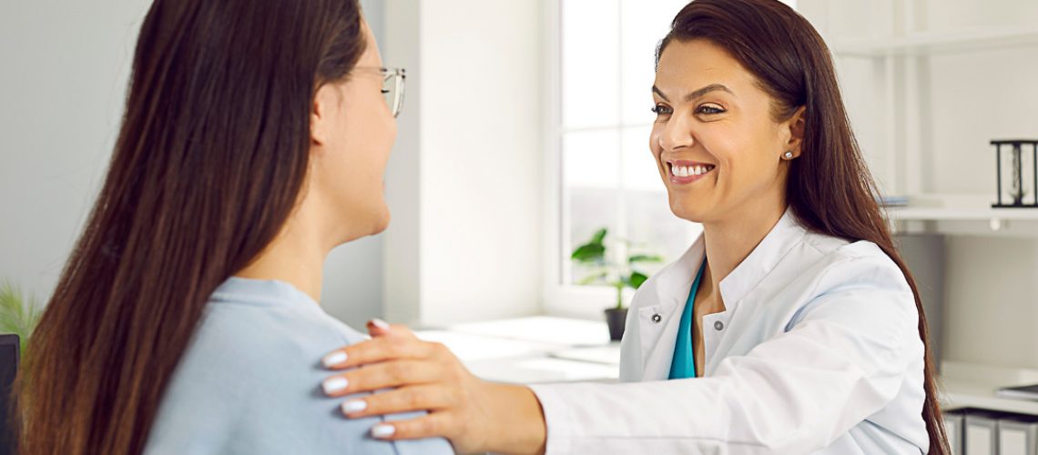 Friendly smiling female doctor holding woman's shoulder telling her positive news about her health. Woman in white coat welcomes female patient in her office. Concept of medicine.