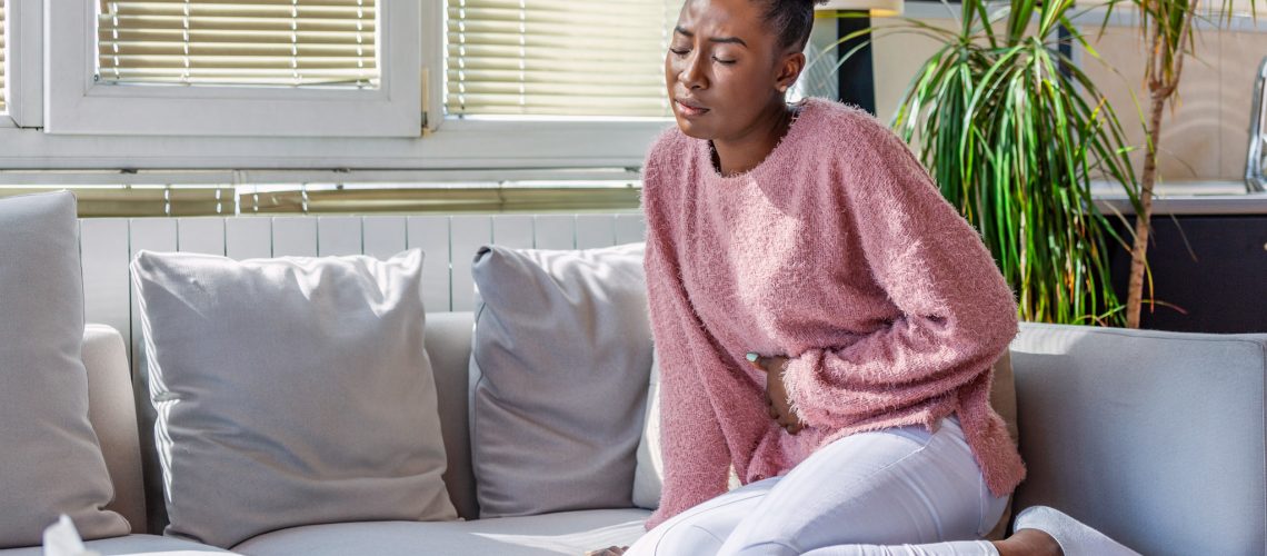 Young black woman suffering from stomachache on sofa at home. Woman sitting on bed and having stomach ache. Young woman suffering from abdominal pain while sitting on sofa at home