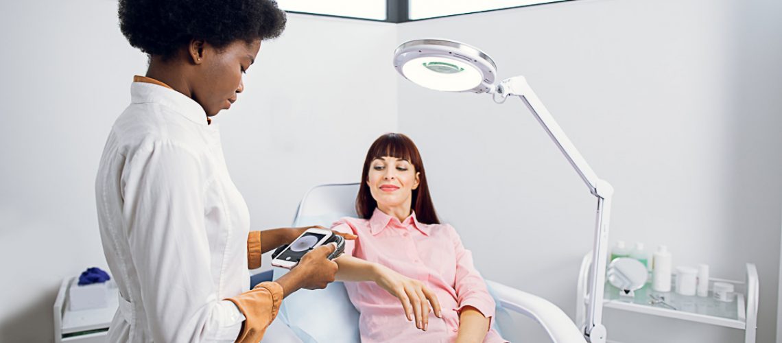 Skin cancer and melanoma prevention. Professional African lady doctor dermatologist examines the patient's moles on arm with the help of a modern device for the dematoscopy